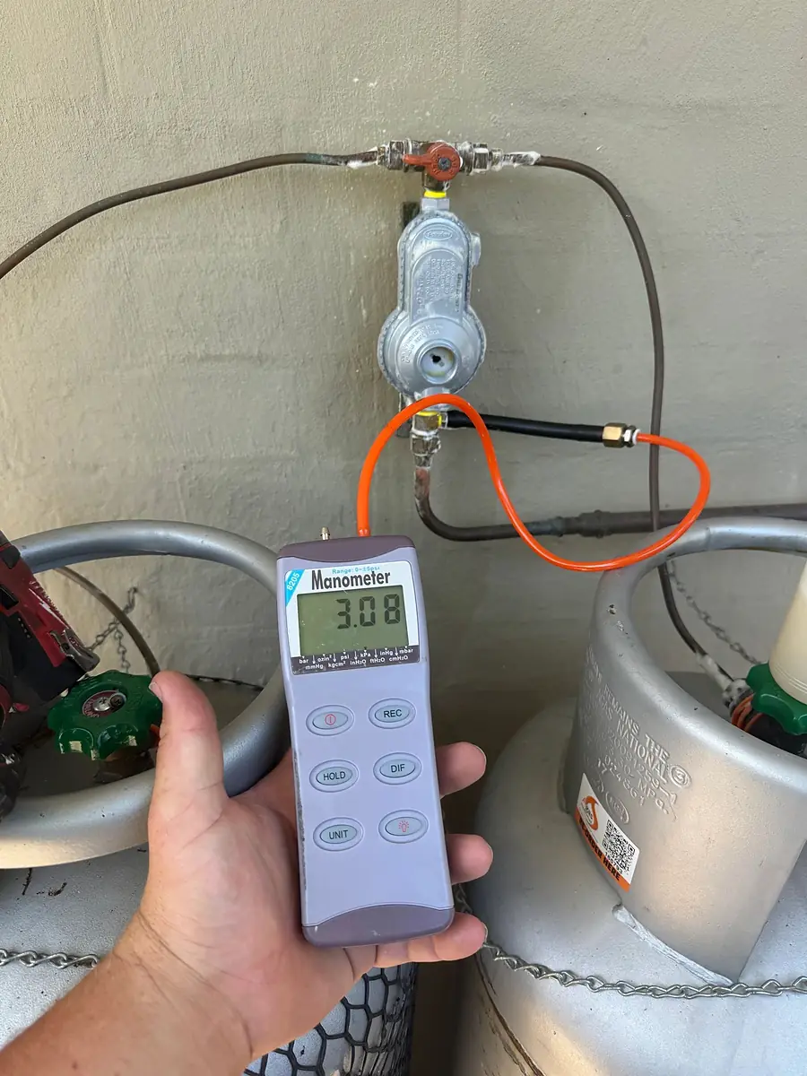 Hand holding a digital manometer reading 3.08 connected to an LPG bottle regulator